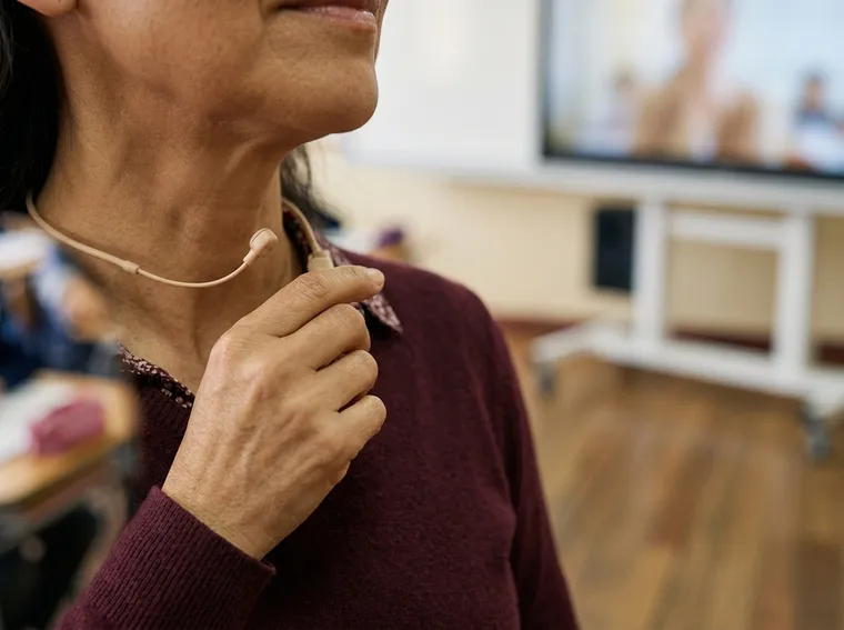 Primer plano de una docente con micrófono de diadema, llevándose la mano al cuello